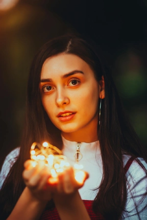 O tânără cu păr lung și închis la culoare ține luminițe aprinse în mâini, privind direct către cameră, pe un fundal întunecat cu iluminare delicată.